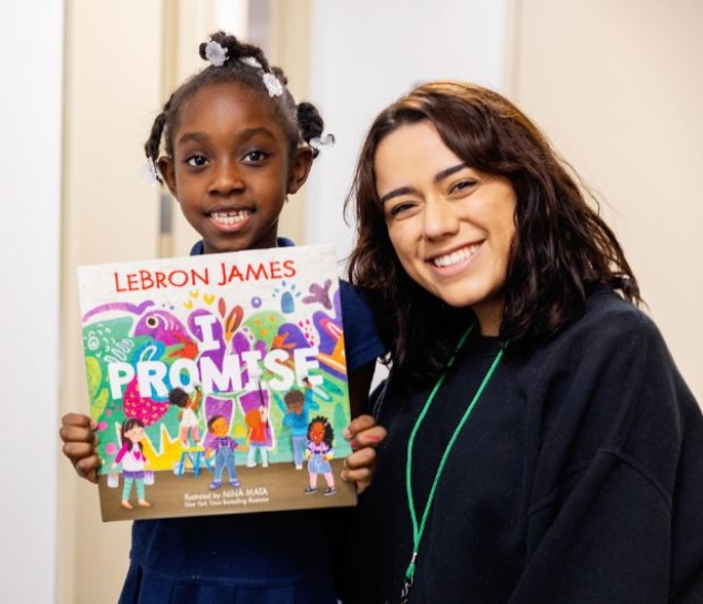 A child holds up the book "I Promise" by LeBron James, standing next to an adult who is also smiling at the camera.