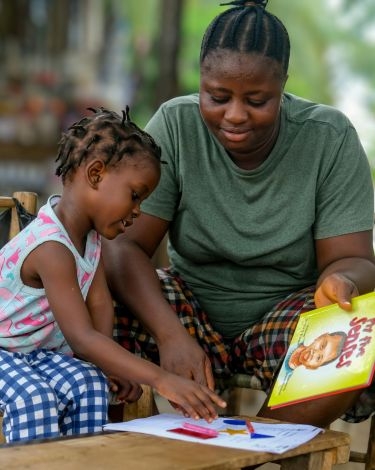 A woman and a child are sitting at a table, looking down at a piece of paper with colorful shapes on it. The woman is holding a yellow book with a picture of a child on the cover.